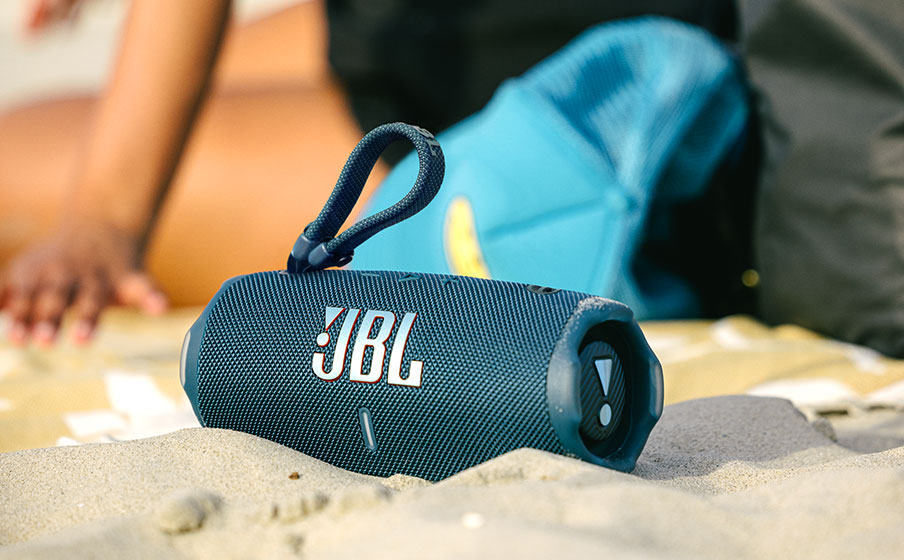 Głośnik JBL Charge 6 gra na plaży obok odpoczywającej osoby. Ilustracja sugeruje długi czas odtwarzania głośnika.