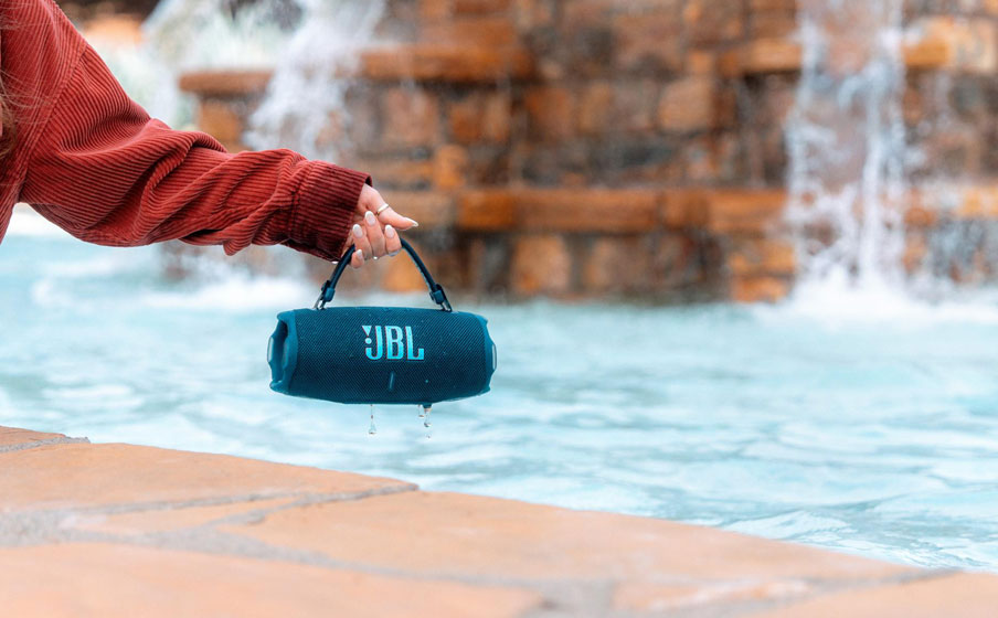 Osoba trzyma głośnik JBL Charge 6 za uchwyt paskowy nad brzegiem fontanny. Ilustracja odporności głośńika JBL Charge 6 na wodę, kurz i upadek.