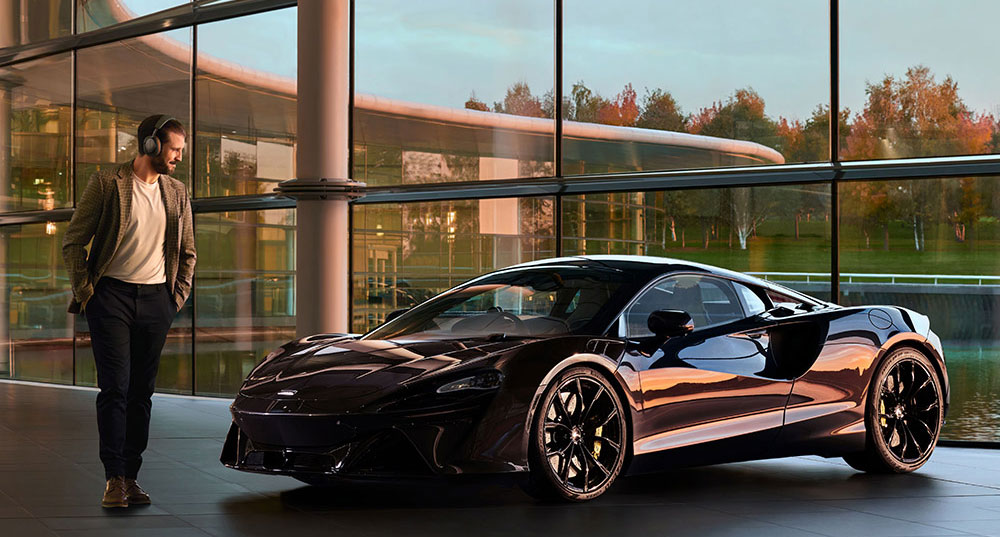 Słuchawki Px8 McLaren opracowano, aby uczcić wieloletnią współpracę obydwu brytyjskich producentów. Zdjęćie przedstawia osobę z założonymi słuchawkami Px8 stojącą obok samochodu McLarena model Artur.