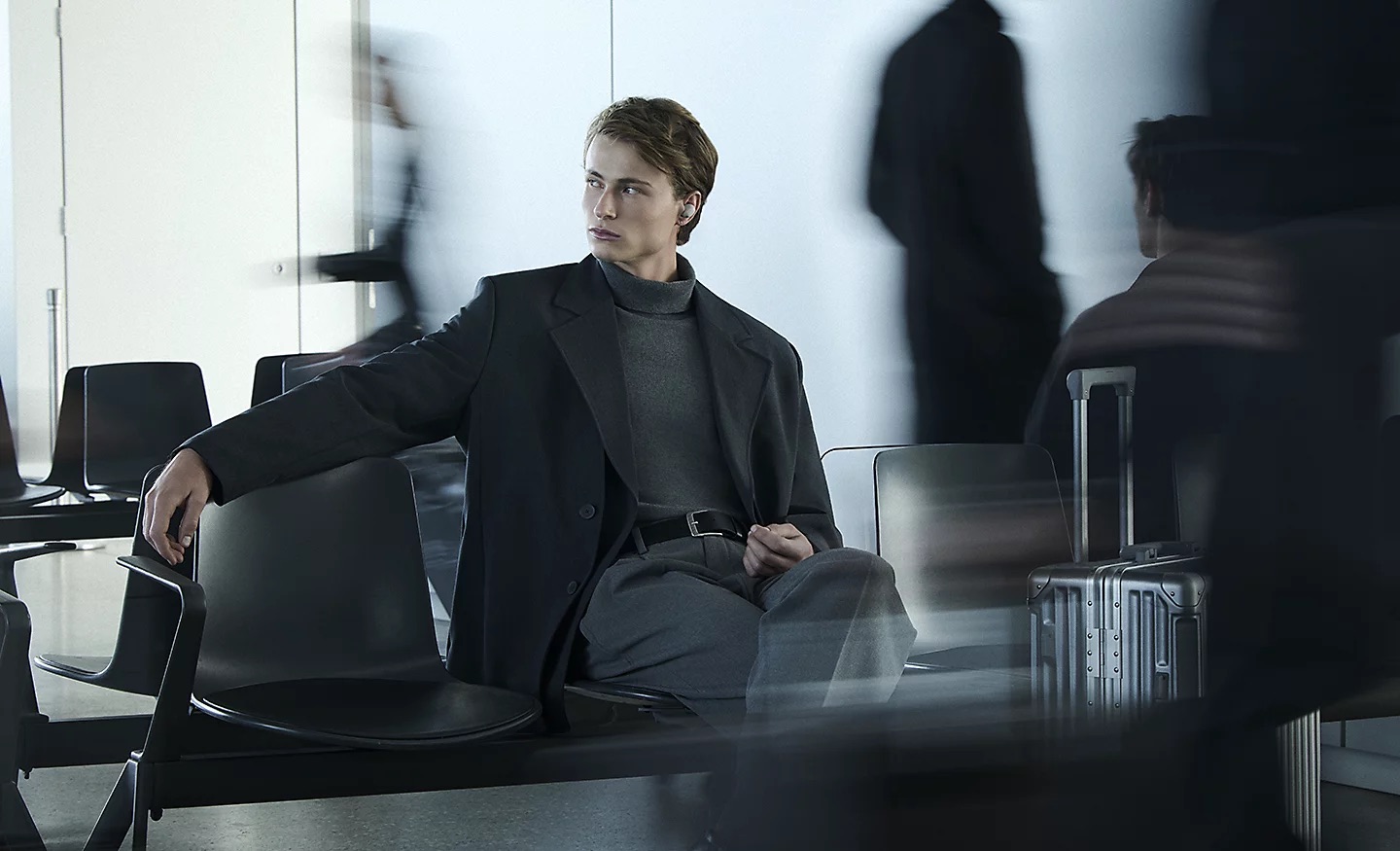 Sony WF-1000XM6 Młody mężczyzna siedzi z walizką w scenerii przypominającej lotnisko. W lewym uchu ma białą słuchawkę, a wokół niego niewyraźne postacie.