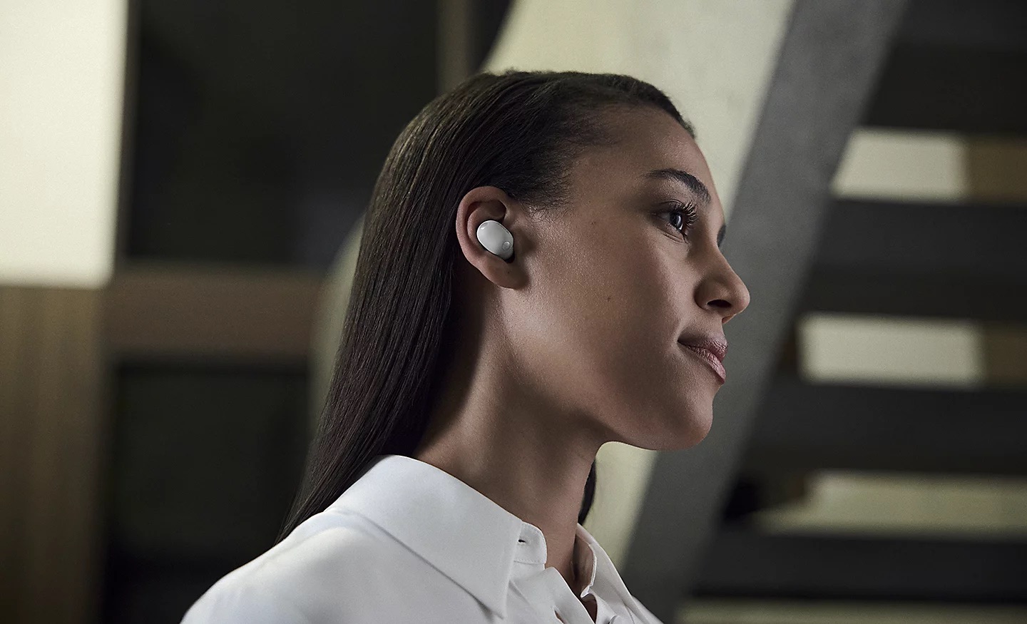 Sony WF-1000XM6 Zdjęcie z profilu kobiety w białej słuchawce dousznej i białej koszuli w nowoczesnym budynku.