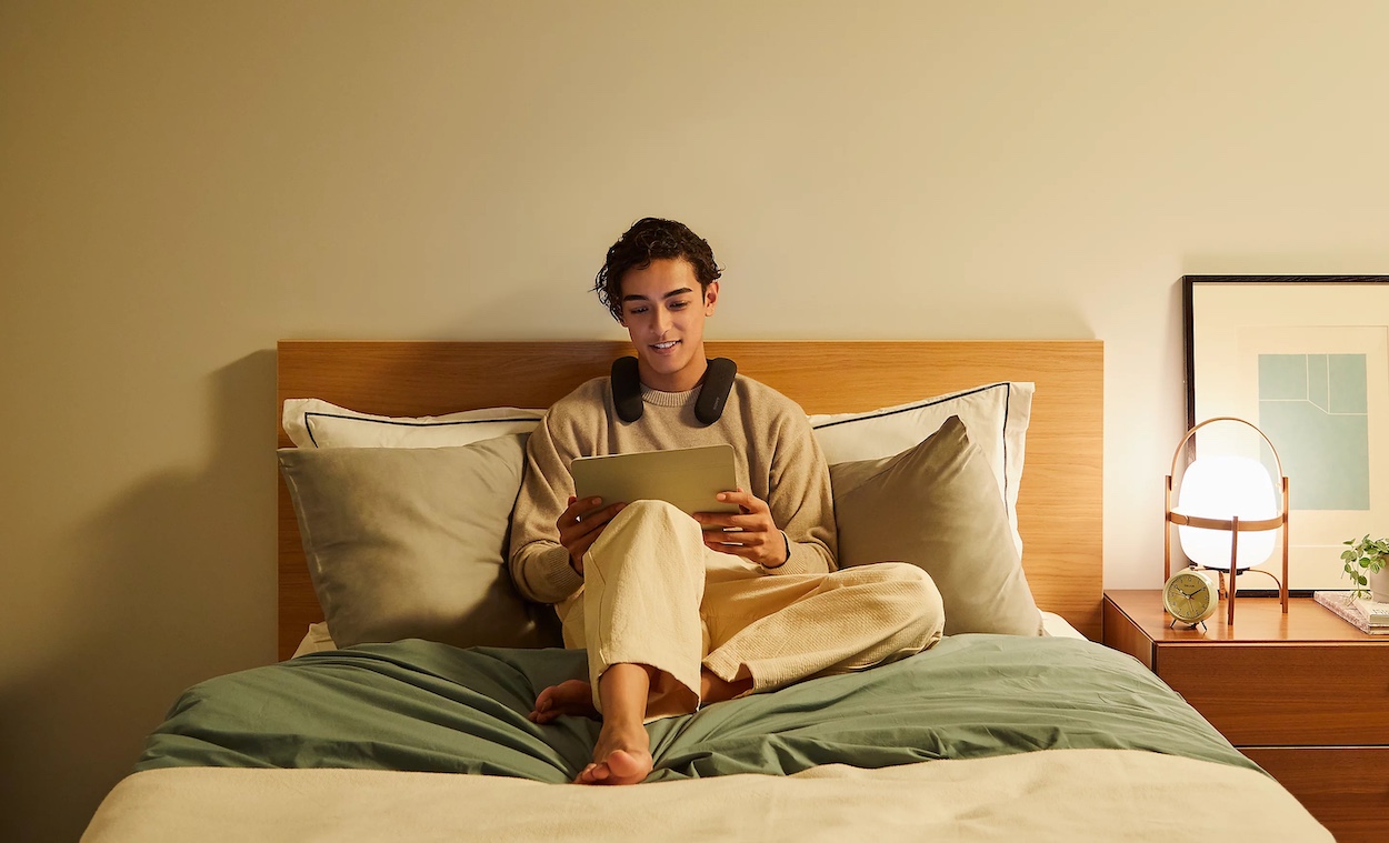 Osoba używająca głośnika BRAVIA Theatre U, siedząca na łóżku i oglądająca coś na tablecie; z prawej stolik nocny z lampką i zegarem