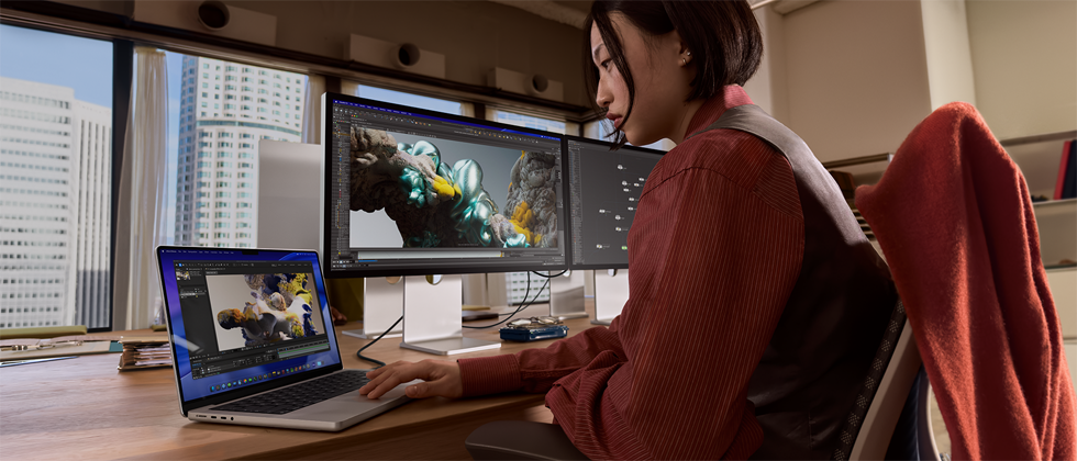 Osoba pracująca w biurze na MacBooku Pro podłączonym do monitora Studio Display XDR, przez okna widać wysokie budynki