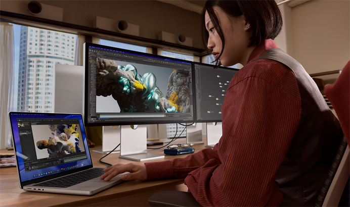 Osoba pracująca w biurze na MacBooku Pro podłączonym do monitora Studio Display XDR, przez okna widać wysokie budynki