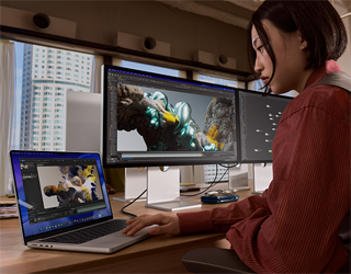 Osoba pracująca w biurze na MacBooku Pro podłączonym do monitora Studio Display XDR, przez okna widać wysokie budynki